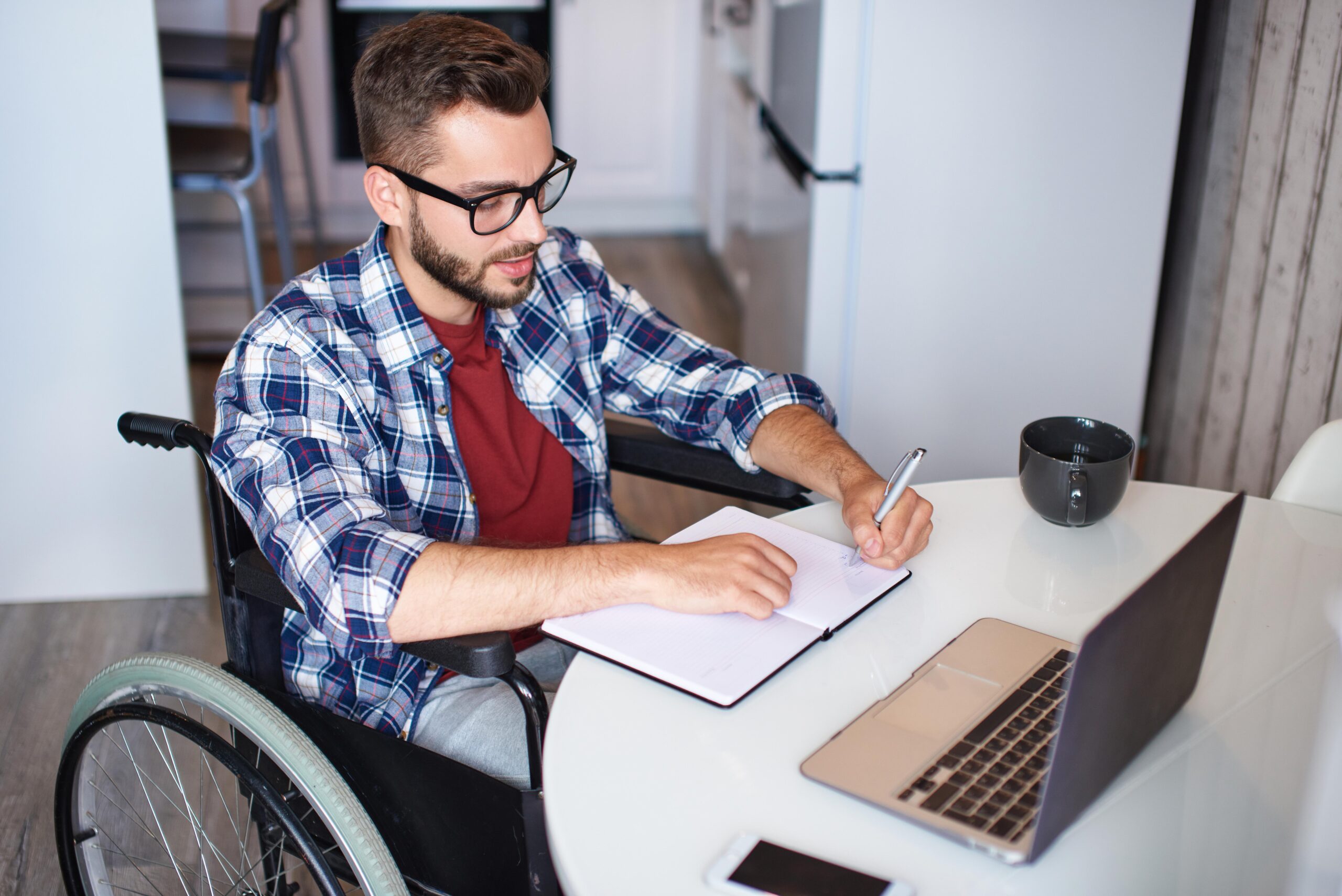 Disabled young man making notes in notepad while using laptop Disabled young man making notes in notepad while using laptop